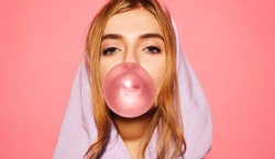 Portrait of young playful blonde girl dressed in blue hoodie standing isolated over pink background while blowing bubble with chewing gum. Woman looking at camera