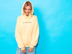 Portrait of young beautiful smiling girl in trendy summer hipster yellow hoodie.Sexy carefree woman posing near blue wall. Positive model having fun.Winks and shows tongue