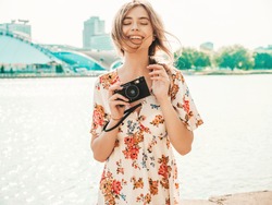 Portrait of young beautiful smiling hipster girl in trendy summer sundress.Sexy carefree woman posing on the sea background at sunset. Positive model holding retro camera