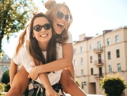 
Two young beautiful smiling hipster female in trendy summer white t-shirt clothes.Sexy carefree women posing on street background. Model jumping on her friend back, gives piggyback riding outdoors