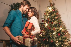 Smiling beautiful woman and her handsome boyfriend. Happy cheerful family posing in the interior near Christmas tree. Models hugging. Love, x-mas concept. They holding gift boxes for each other
