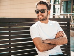 Portrait of handsome smiling stylish hipster lambersexual model.Man dressed in white T-shirt. Fashion male sitting at the bench in the street background in sunglasses. Crossed arms