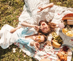Two young beautiful smiling hipster female in trendy summer sundress and hats.Carefree women making picnic outside.Positive models sitting on plaid on grass, hugging, eating fruits and cheese
