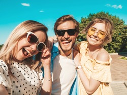 Group of young three stylish friends in the street.Man and two cute girls dressed in casual summer clothes.Smiling models having fun in sunglasses.Women and guy making photo Pov selfie on smartphone