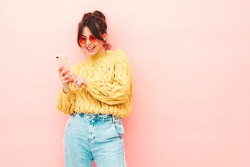Young beautiful smiling female in trendy summer yellow hipster sweater and jeans.Sexy carefree woman posing near pink wall in studio.Positive model having fun. Looking at smartphone screen, using app