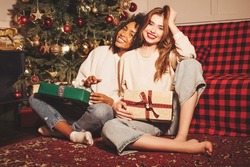 Two young beautiful smiling international hipster female.Sexy carefree women sitting near Christmas tree.Positive models hugging and giving each other gift boxes.Christmas, x-mas, concept