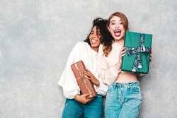 Two young beautiful smiling international hipster female in trendy clothes.Sexy carefree women posing near gray wall.Positive models hugging and giving each other gift boxes.Christmas, x-mas, concept