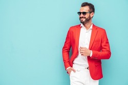 Portrait of handsome smiling stylish hipster lambersexual model.Sexy modern man dressed in elegant red suit. Fashion male posing in studio near blue wall in sunglasses