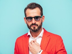 Portrait of handsome confident stylish hipster lambersexual model.Sexy modern man dressed in elegant red suit. Fashion male posing in studio near blue wall in sunglasses