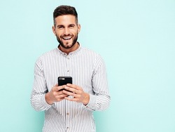 Handsome smiling model.Sexy stylish man dressed in shirt and jeans. Fashion hipster male posing near blue wall in studio. Holding smartphone. Looking at cellphone screen. Using apps