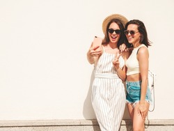 Two young beautiful smiling hipster female in trendy summer clothes.Sexy carefree women posing in the street near white wall in hat. Positive pure models having fun at sunset, taking selfie