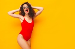 Young beautiful smiling woman posing near yellow wall in studio.Sexy model in red swimwear bathing suit.Positive female with curls hairstyle.Happy and cheerful. In sunglasses