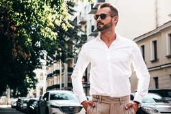 Portrait of handsome confident stylish hipster lambersexual model.Sexy modern man dressed in white shirt and trousers. Fashion male posing on street background in Europe city at sunset. In sunglasses