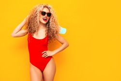 Young beautiful smiling woman posing near yellow wall in studio.Sexy model in red swimwear bathing suit.Positive female with afro curls hairstyle. Holding penny skateboard. Happy and cheerful