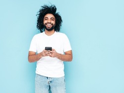 Handsome smiling hipster  model.Sexy Arabian man in summer stylish clothes.Fashion male with long curly hairstyle posing near blue wall in studio.Looking at cellphone screen. Using apps