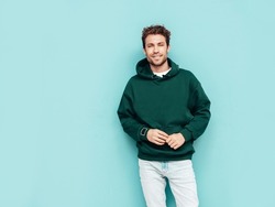 Handsome confident hipster  model.Sexy unshaven man dressed in summer stylish green hoodie and jeans clothes. Fashion male with curly hairstyle posing in studio. Isolated on blue