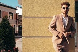 Handsome confident stylish hipster lambersexual model.Sexy modern man dressed in elegant beige suit. Fashion male posing in the street background in Europe city at sunset. In sunglasses