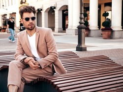 Handsome confident stylish hipster lambersexual model.Sexy modern man dressed in elegant beige suit. Fashion male posing in the street in Europe city at sunset. In sunglasses. Sitting at the bench