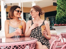 Two young beautiful smiling hipster women in trendy summer dress clothes.Sexy carefree models sitting in terrace cafe.Positive female eating tasty ice cream in waffles cone at sunny day. In sunglasses