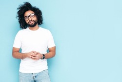 Handsome smiling hipster  model.Sexy unshaven Arabian man dressed in white summer t-shirt and jeans clothes. Fashion male with long curly hairstyle posing near blue wall in studio. In glasses