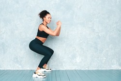 Fitness smiling black woman in sports clothing with afro curls hairstyle.Wearing sportswear.Sexy young beautiful model doing lunges before training. Female making squats near grey wall