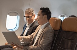 Two passengers looking at laptop together on board. Businessman in brown suit showing project on notebook to businessman with eyeglasses. Work and travel concept.