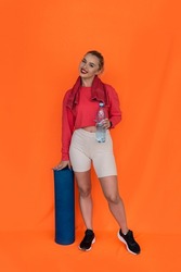 a beautiful young girl in a top and shorts holds a mat twisted into a tube in her hands on a plain background. The concept of a sporty girl with a mat on the background