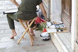 Woman artist painting on the street, she is sitting on a wooden bench seat, tools paint beside her feet