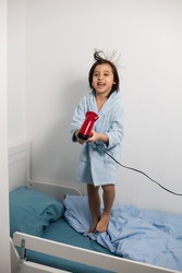 five year old brunette boy with long hair and in blue robe dries his hair with a red hair dryer ,standing on the bed in the children's room
