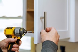 The worker sets a new handle on the white cabinet with a screwdriver installing kitchen cabinets