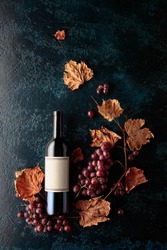 Bottle of red wine with ripe grapes and dried up vine leaves. Old dark blue background. On a bottle old empty label. Copy space, top view.