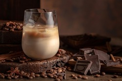 Irish cream and coffee cocktail in a glass with ice on an old wooden background. Coffee beans, cinnamon, anise, and pieces of chocolate are scattered on the table. 