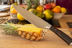 Close-up view of a damascus kitchen knife in half a pineapple fruit on a cutting board in a domestic kitchen with other varieties of fruit in the background