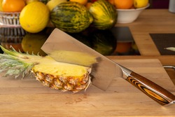 Close-up view of a cleaver in half a pineapple fruiton a cutting board in a domestic kitchen with other varieties of fruit in the background