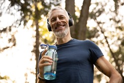 Low angle portrait of joyful grizzled athletic male drinking water while doing workout in sunny forest