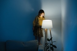 Brunette little girl in yellow knitted sweater and leopard skirt standing on a backseat of a couch at home. She's looking at dim night light from above. Her face is lit.