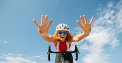 Low angle shot of vigorous hyper-emotional young ginger woman riding on a professional bike hands free, showing them to camera. She's geared up, wearing helmet and cycling jersey.