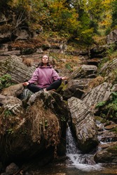 Young woman sitting barefoot on a big stone cross-legged, meditating. Over mountain spring with huge square boulders and autumn deciduous trees above.