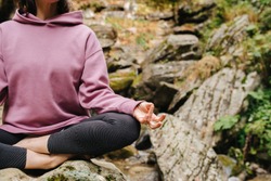 Young woman sitting barefoot on a big stone cross-legged, meditating. Over mountain spring with huge square boulders. Close up, cropped, no face.