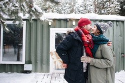 Confident man in winter clothes kissing his woman, while hiding the present behind his back. Standing in front of a small snowy house.