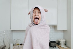 Happy funny little girl wrapped in a bear hooded towel sticking tongue out, her eyes closed.