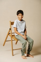 Amused indian boy sitting on a wooden folding chair in a beige room. He is leaning forward with the chair, looking at the camera.