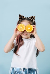 Funny tween girl with cat ears holding orange halves over her eyes. She has dyed hair strands. Posing over blue background.
