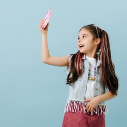 Fancy tween girl in jeans jacket, skirt taking a selfie, looking up. She has partially dyed hair strands. Posing over blue background.