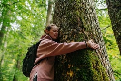 Upward view of a peaceful young woman hugging a thick moss grown tree in a forrest. She is wearing pink hoodie.