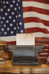 Old wooden desk with vintage typewriter holding an empty sheet of paper in front of the USA flag
