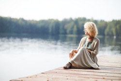 Serious pensive curly-haired mature lady in long skirt sitting on pier and contemplating tranquil nature around