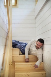 Young Caucasian man in homewear lying on wooden staircase at his house and wincing at pain after falling from high distance