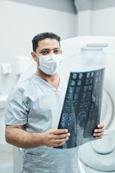 Professional mixed-race radiologist in mask and uniform holding x-ray image of patient head against environment of medical office
