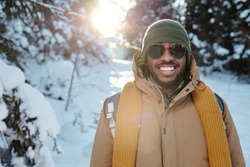 Happy young African man in sunglasses and warm winterwear standing in front of camera against firtrees in snow during chill on winter weekend